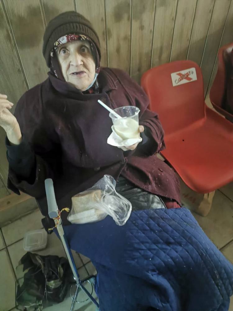 Femeie în vârstă, cu căciulă, care mănâncă o porție caldă, așezată lângă un scaun roșu
