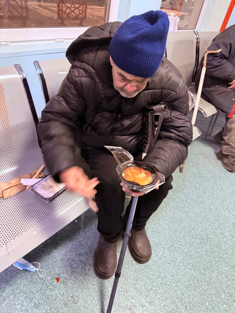 Bărbat în vârstă, cu fes albastru, care mănâncă o porție caldă pe o bancă din gară
