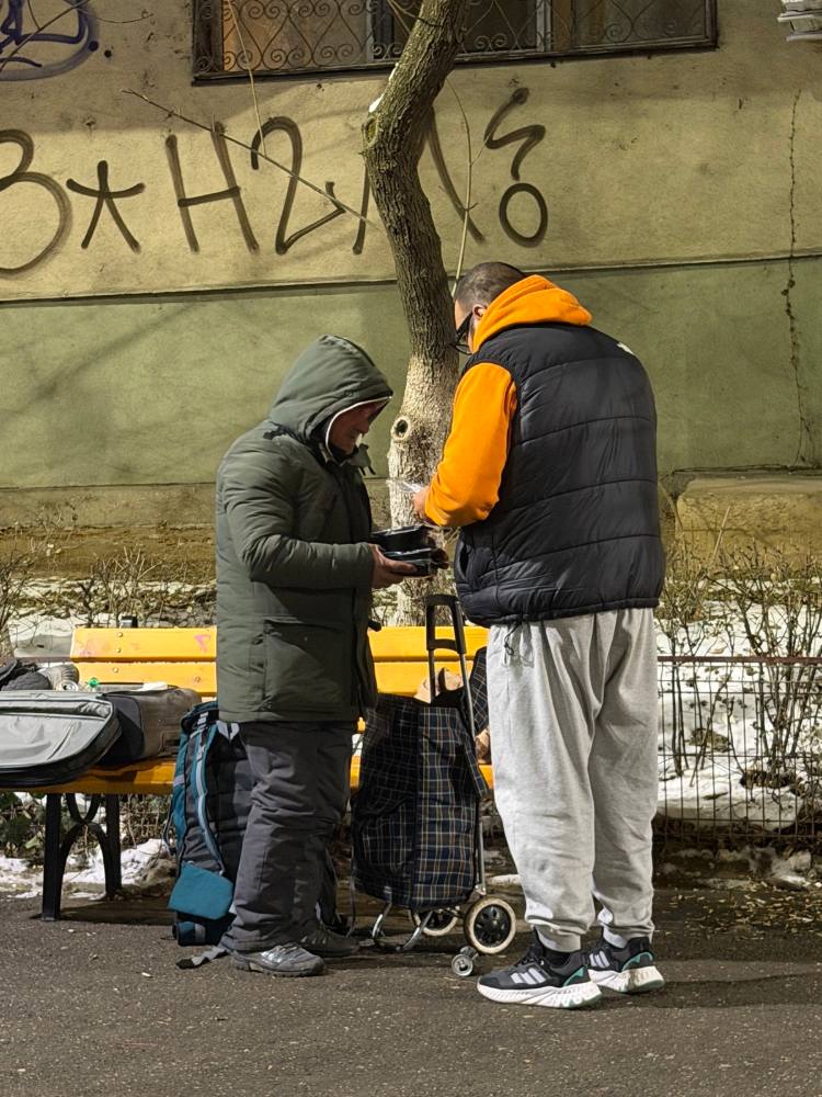 Voluntar care dă mâncare unui om pe stradă, iarna, lângă o bancă